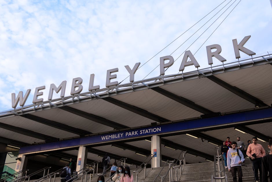 The exterior of Wembley Park Station in London with large, white, three-dimensional letters spelling 'WEMBLEY PARK' mounted on the station's roof structure against a partly cloudy sky. Below the sign, a blue panel displays the station name 'WEMBLEY PARK STATION.' The station entrance features a set of stairs with metal handrails, and several passengers, some wearing face masks, are seen ascending and descending. The station's architecture includes a flat, overhanging roof supported by dark metal beams. The scene captures a busy, urban transportation hub typical of a London underground station, relevant to house removals and moving services involving transportation logistics near Wembley Park.
