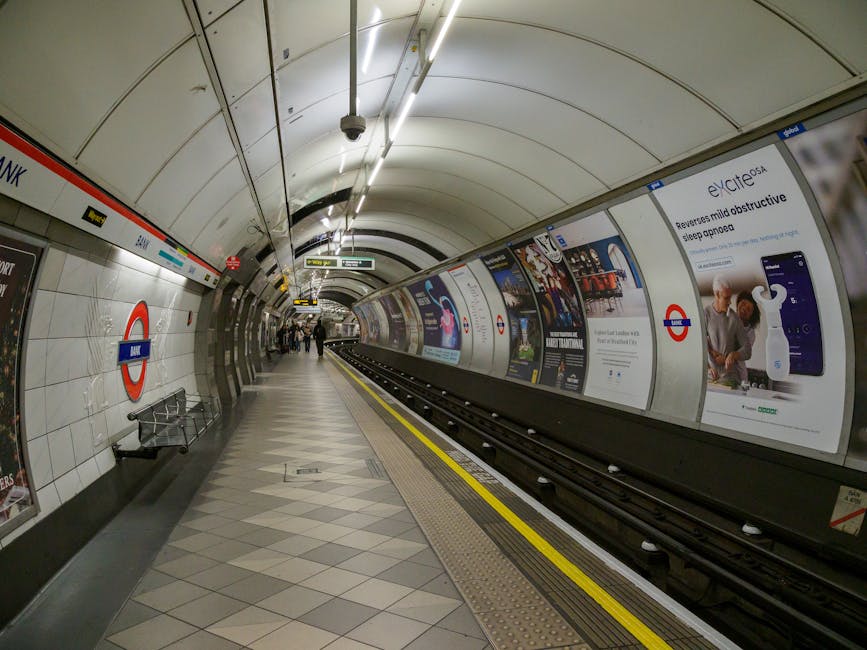 Inside the underground station platform near East Finchley tube station in Finchley, London, the image shows a curved platform with white tiled walls featuring the London Underground roundel sign and several advertising posters along the wall. The platform has a patterned tile floor with grey and beige tiles, and a yellow line demarcates the safe waiting area next to the tracks. Overhead, bright fluorescent lighting illuminates the space, and a few commuters are visible in the distance, walking or standing. The station's tunnel ceiling curves overhead, and there are metal railings and benches for passengers. The platform provides a clean, modern environment typical for a busy London transportation hub, supporting the process of home relocation or furniture transport by facilitating efficient movement and accommodation of moving vehicles or staff during packing and loading processes. Occasionally, the station is cleaned or maintained, and the scene captures a typical moment during daily operations, with Movers Finchley possibly involved in logistics near this area.