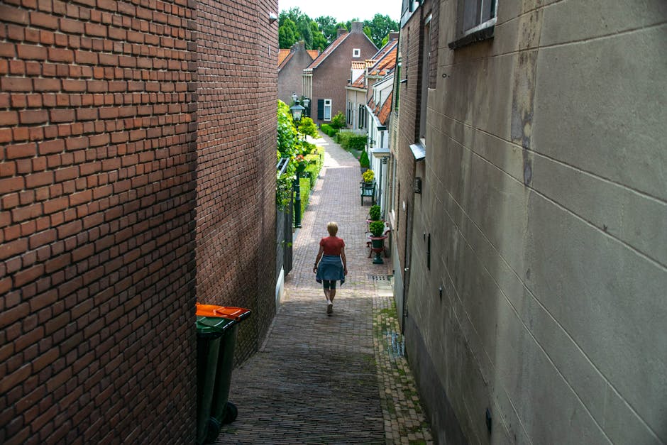 A narrow cobblestone street in a residential area with red-brick houses featuring black-framed windows and sloped roofs. In the foreground, a brick wall runs along the left side of the street, partial