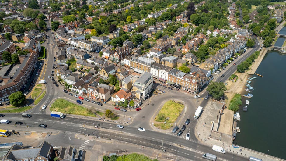 An aerial view of North Finchley Tally Ho area showing a variety of buildings including residential houses and commercial properties along the streets, with a river on the right side featuring boats docked along the promenade. In the foreground, a loading area adjacent to a street is visible where Movers Finchley is involved in home relocation activities. The area includes parked cars, delivery trucks, and a small paved space with patches of grass, with some furniture and boxes likely being prepared for transport. The scene highlights the urban setting with clear roads, greenery, and the movement of vehicles during a busy day of furniture transport and packing in the context of house removals in the North Finchley area.