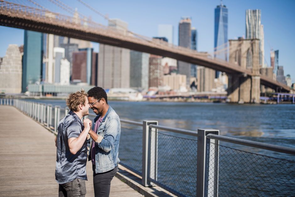 Two men standing on a wooden pier beside a river in an urban area, with the Brooklyn Bridge visible in the background and a city skyline of tall skyscrapers. One man has curly blond hair and is wearing a short-sleeved shirt, while the other has short dark hair, glasses, and is dressed in a denim jacket. They are smiling and holding hands, appearing to share a joyful moment. The scene is under bright daylight with clear skies and calm river water. Although the image features a pair of people, it is related to the context of city life and relocation services in North Finchley, with Movers Finchley operating in the removals sector, potentially supporting home relocation or furniture transport amid the urban environment.